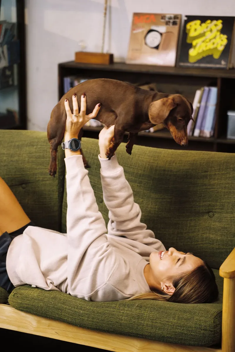 Person lying on a green sofa lifting a dachshund overhead