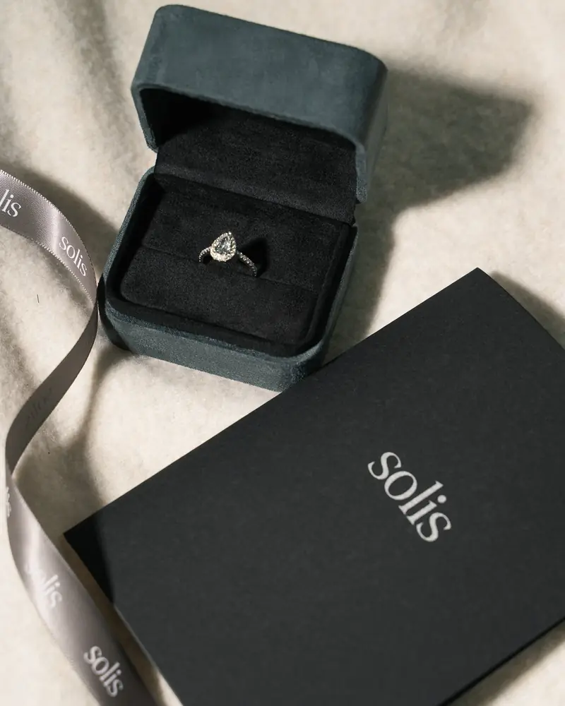 Engagement ring in an open dark-green box beside a Solis gift box