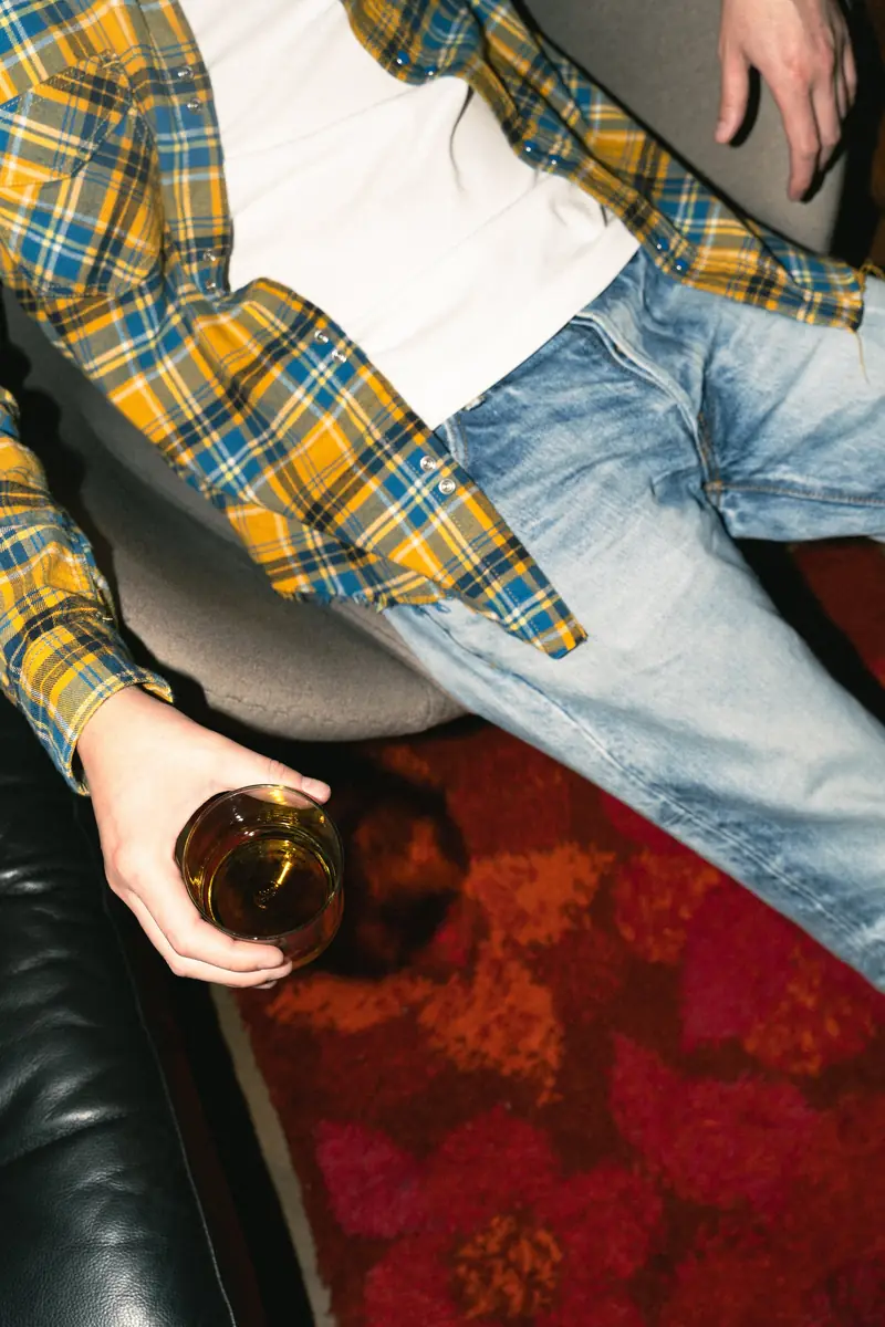 Person in a checked shirt and jeans holding a bottle on a leather sofa